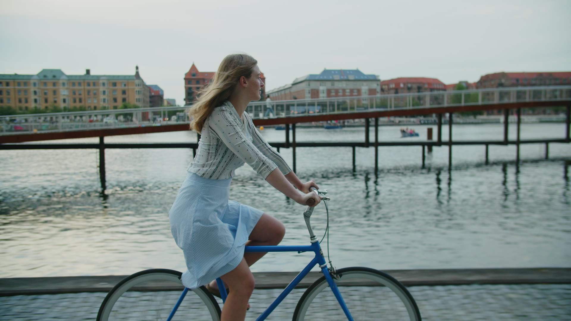 En kvinde cykler langs kajen på Islands Brygge i det flade lys efter solnedgang, med havnebadets bro bag sig.